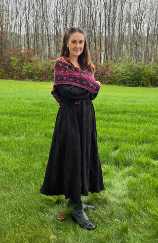 A woman stands in a grassy field wearing a Season of the Witch 2026 Shawl pattern, featuring dark grey and magenta gothic lace over a long black dress.