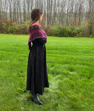 A person wearing a black and bright pink patterned Season of the Witch 2026 Shawl Pattern stands in a grassy field in a long black dress.