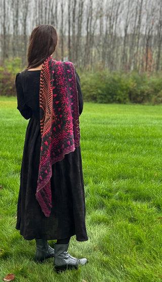 A person seen from behind wears a black dress and a long, asymmetrical Season of the Witch 2026 Shawl Pattern featuring vibrant shades of orange, purple, and black in a geometric pattern.