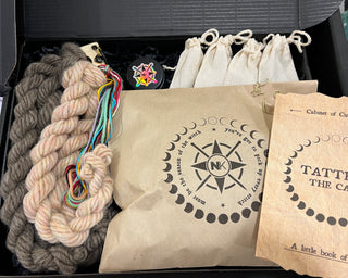 A Tatters the Cat Poppet | Family Knitting Kit in a black box containing two hanks of yarn, several small drawstring pouches, a black jar with a circular sticker, a cat-shaped stitch marker, and a printed booklet.