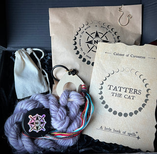A black box containing a Tatters the Cat Poppet | Knitting Kit, featuring a skein of purple yarn, a wooden tool, a small drawstring bag, and an instruction booklet.
