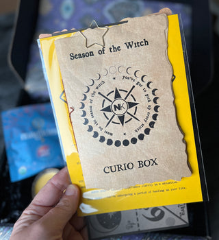 A hand holds a Knitter’s Curio Box information card decorated with a compass graphic and moon phases, secured with a star-shaped paperclip.