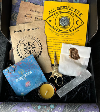 An open Knitter’s Curio Box containing a yellow fortune card, a lunar-designed flyer, a tea packet, a small candle, metallic stork-shaped scissors, a wax-sealed envelope, and a selenite wand.
