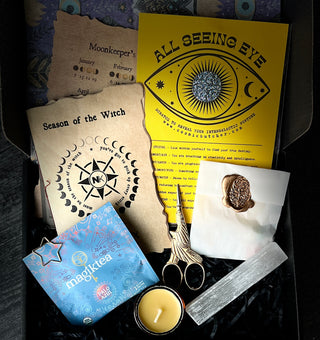 An open Knitter’s Curio Box filled with craft supplies, including a yellow All Seeing Eye card, a paper project, a tea bag, gold bird-shaped scissors, a small candle, and a selenite wand.