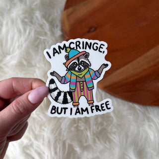 Sticker with a raccoon illustration and text that says I am cringe but I am free held by a hand over a wooden surface.