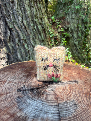 A small, rectangular Tatters the Cat Poppet with embroidered features and a small flower, resting on a tree stump.