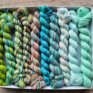 A Koigu Pencil Box in the Osheaga colorway, featuring neatly arranged mini skeins of Merino Wool Fingering Sock Yarn, blends green, yellow, turquoise, and mint hues as it rests on a wooden surface.