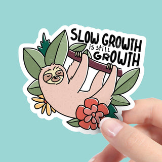 Sticker with a sloth holding a branch with 'Slow Growth is Still Growth' text, held by a hand against a light blue background.