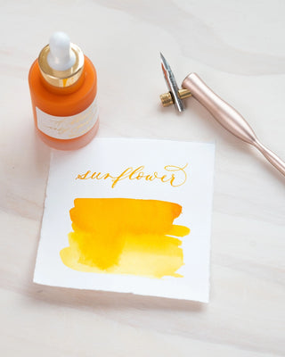 A bottle of Premium Calligraphy Ink, a metallic pen, and a sheet featuring “sunflower” in gold script with yellow watercolor swatches are arranged on a light wooden surface.