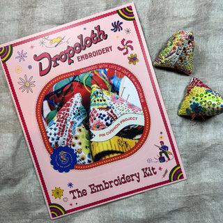 The Hand-Embroidered Pin Cushion Project Kit, shown on fabric with two finished, colorful cushions, features decorative designs and text on the packaging and includes a tutorial for making your own hand-embroidered pin cushions.