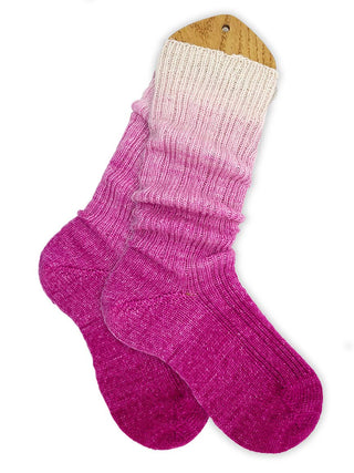 Two gradient socks made from Solemate Sock Yarn are displayed on a wooden blocker, featuring a beautiful fade from light pink at the cuff to deep magenta at the toes and heels.