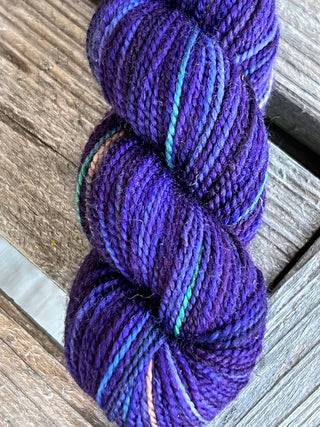 A skein from the KPM and KPPPM Variegated Color Collection, in deep purple with blue and turquoise streaks, rests on a weathered wooden surface—perfect for knitting cozy projects.