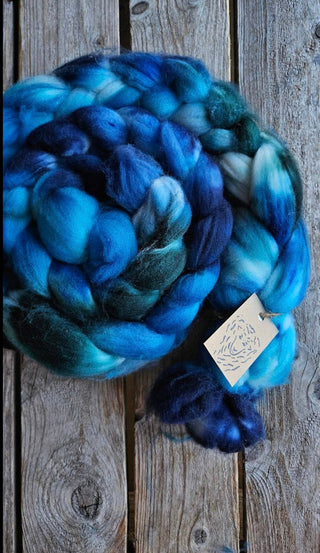 A thick braid of Koigu Merino Roving in blue, teal, and white rests on a rustic wooden surface, a tag attached to this soft spinning fiber.