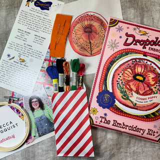 A flat lay of the Cosmic Poppy Embroidery Kit, showing colorful threads, a pink kit box, floral poppy sampler fabric, needle, instructions, and a smiling woman in overalls.