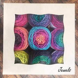 A square box with a wavy-edged clear window displays nine colorful Freia Minikin Palette Packs yarn balls in the "Jewels" color, arranged in a grid—ideal for Fair Isle sweaters. "Jewels" is written in cursive on the bottom right corner.