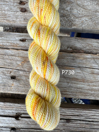 A twisted skein of KOIGU Bridgerton Collection KPPPM hand knitting yarn in color P730 (yellow, cream, and orange) rests on a weathered wooden surface with the label overlaid near this premium merino wool skein.