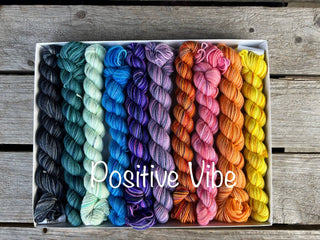 The Spooky Season Koigu Pencil Box in Positive Vibes features merino wool mini skeins arranged in a vibrant gradient from black to yellow—perfect for adding color to your next project. Box shown on a wooden surface.