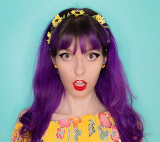 A person with long purple hair, blunt bangs, and a sunflower headband wears handmade 🌻 Sunflower Earrings 🌻 and a yellow floral top, standing against a bright blue background with a surprised expression.