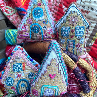 The Gingerbread Houses Embroidered Ornaments Sampler includes three hand-embroidered fabric ornaments shaped like gingerbread houses, featuring candy, hearts, and windows in vibrant thread against a quilted background.