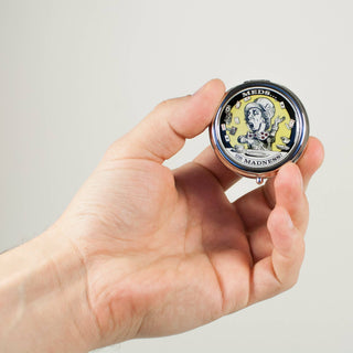 A hand holds the Mad Hatter Stitch Marker / Pill Box, a small round container with a label featuring a distressed cartoon figure surrounded by pills and text "MEDS. ON MADNESS," set against a plain light-colored background.