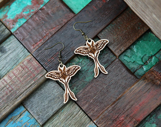The Luna Moth Maple Wood Earrings, crafted with detailed wing and body accents from maple wood, are displayed on a textured wooden surface—a unique accessory for gothic earring enthusiasts.