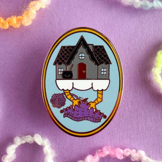 The Baba Yaga Knitting Enamel Pin features a chicken-legged house on a purple surface with pins and yarn, against a light blue background. A cute addition to any collection—ideal for knitting pin enthusiasts!.