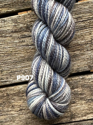 A skein from the KPM and KPPPM Variegated Color Collection in grey, blue, and brown rests on weathered wood. Ideal for hand knitting, the soft merino yarn features label P907 printed in white next to it.