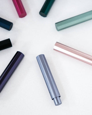 Several colorful The Wren Refillable Writing Pens are arranged in a circle on a white surface, their ends pointing outward. Each eco-friendly pen features a unique metallic color such as silver, pink, green, and purple.