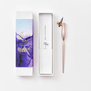 The Flourish Calligraphy Pen, featuring a metallic pink holder and nib, sits beside an open white and purple decorative box adorned with a palm tree illustration and cursive writing inside.