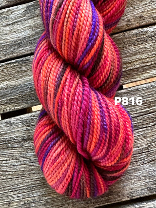 A KPM and KPPPM Variegated Color Collection Skein in red, pink, orange, and purple rests on rustic wood. Perfect for hand knitting projects. The image features the label P816.