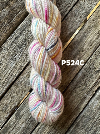 A skein from the KPM and KPPPM Variegated Color Collection rests on rustic wood, showcasing pastel hues with specks of pink, yellow, blue, and black. The code P524C is displayed beside this soft merino wool, ideal for hand knitting.