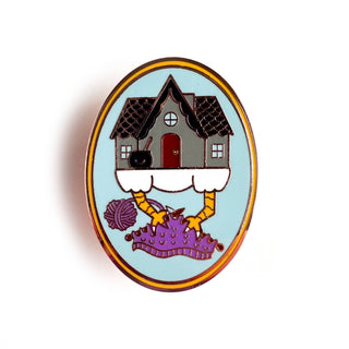 The Baba Yaga Knitting Enamel Pin features a charming chicken-legged house atop a purple crown, with yarn balls and knitting needles below, set on a light blue background and framed by a thin yellow border.
