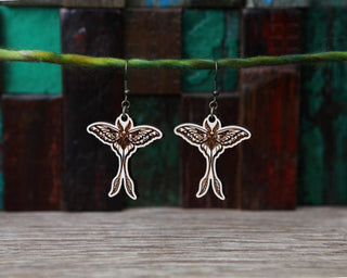 A pair of Luna Moth Maple Wood Earrings featuring intricate wing and body designs hang from a green string against a background of colorful wooden blocks.