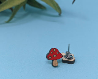 The Mushroom Earrings, hypoallergenic and handmade in the USA, are small stud earrings shaped like red and white spotted mushrooms. Displayed on a light blue surface with a blurred green plant in the background.
