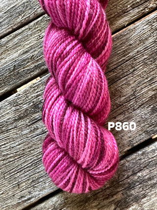 A skein from the KPM and KPPPM Variegated Color Collection in pink and magenta rests on a weathered wooden surface. Neatly twisted merino wool, ideal for hand knitting, features a label marked “P860” in white text.