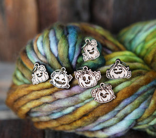 Five 🎃 Pumpkin Jack O’Lantern Stitch Markers with smiling faces rest on a skein of multicolored yarn in green, brown, and purple tones, shown against a rustic wooden background.