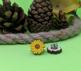 The 🌻 Sunflower Earrings 🌻 are displayed on a green surface, with pine cones, a rope, and leaves artistically arranged in the background.