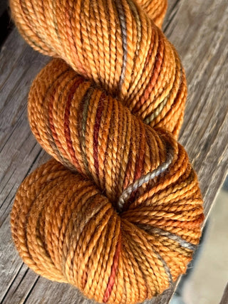 A close-up of a KPM and KPPPM Variegated Color Collection Skein in orange with brown, gray, and red tones, perfect for hand knitting, resting on a weathered wooden surface.