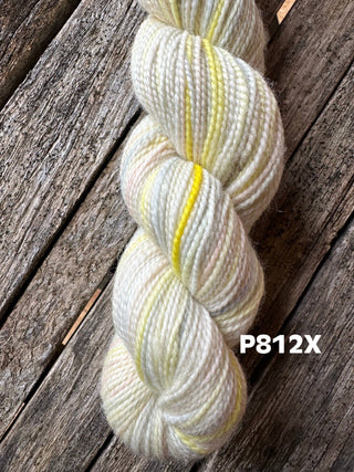 A skein from the KPM and KPPPM Variegated Color Collection rests on weathered wood, featuring white with subtle yellow and pastel streaks. "P812X" appears in white at the lower right—ideal for hand knitting projects.