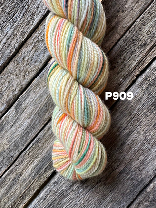 A skein from the KPM and KPPPM Variegated Color Collection rests on a rustic wooden surface, featuring pastel shades of yellow, orange, green, and pink. The label P909 is shown beside this soft merino wool yarn, ideal for hand knitting.