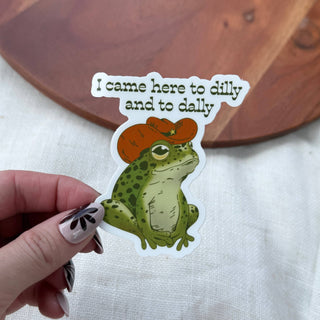 A hand displays the I Came Here To Dilly and To Dally Southern Frog Sticker, a waterproof decal with a green frog in a red cowboy hat and fun text above. The background features white fabric and a wooden surface.