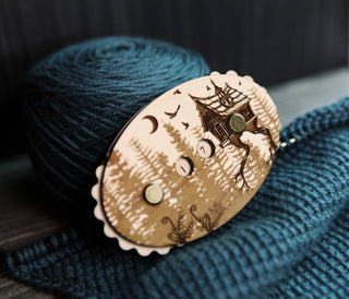 The Baba Yaga Row Counter—an oval wooden pin engraved with a house on bird legs, trees, birds, and a crescent moon in the style of folklore—rests atop a ball of teal yarn and knitted fabric.