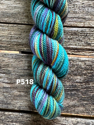 A KPM and KPPPM Variegated Color Collection skein in teal, blue, green, brown, and yellow rests on rustic wood. Ideal for hand knitting projects. The label P518 is shown beside the yarn.