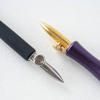 Two One-Dip-Nib calligraphy nibs rest on a white surface: one has a silver nib with a black handle for line variation; the other features an adjustable gold-tipped nib, purple handle, and small gold screw for secure placement.