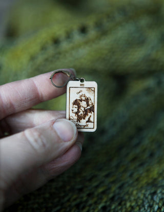 A hand holds a rectangular keychain from the "Tarot Card Stitch Markers – The Hermit, The Magician & Strength" set, featuring the Strength card with a lion, against a green knitted background—perfect as a unique knitting accessory.