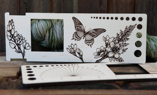 The Butterfly "Almost Everything" Ruler is a wooden knitting/crochet tool featuring floral and butterfly designs, needle size holes, a yarn weight gauge showing a skein of green yarn, and another similar fiber arts ruler placed in front.
