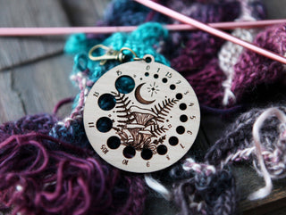 The 🌙 Night Sky & Forest Floor Knitting Needle Gauge 🌙, made of cherry wood with an engraved forest and moon design, sits on vibrant yarn and needles. It features numbered holes to measure knitting needle sizes.