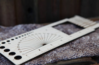A close-up of the Luna Moth “Almost Everything” Ruler, featuring holes and measurement lines, resting on textured knitted fabric. This versatile fiber-arts tool is shown against a softly blurred background.
