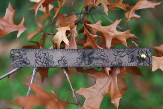 The Oak Leaves & Acorn Leather Ruler Cuff features black sketches of oak leaves and acorns on brown leather, inspired by autumn branches—perfect for those seeking a unique, nature-themed accessory.