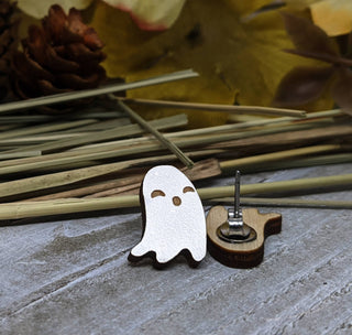 Our handmade Ghost Earrings feature a small wooden ghost shape with a painted white face and hypoallergenic metal backing, displayed on a gray surface with pinecones, dried grass, and autumn leaves.